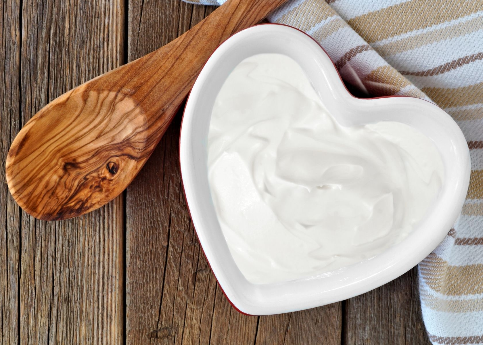 Greek yogurt in white heart-shaped bowl next to wooden spoon and kitchen towel.