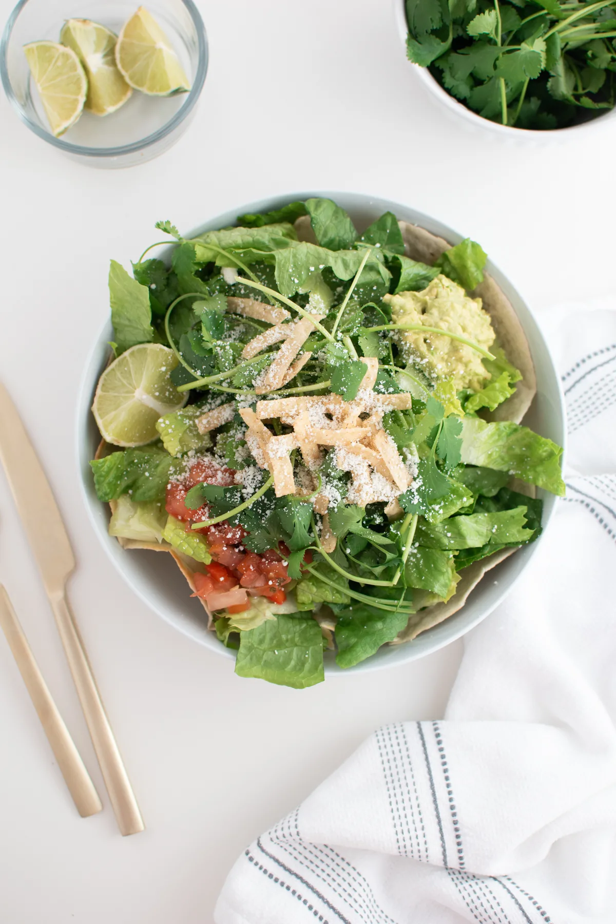 Cafe Rio salad recipe in white bowl surrounded by bowl of limes, cilantro, a white kitchen towel, and gold utensils.