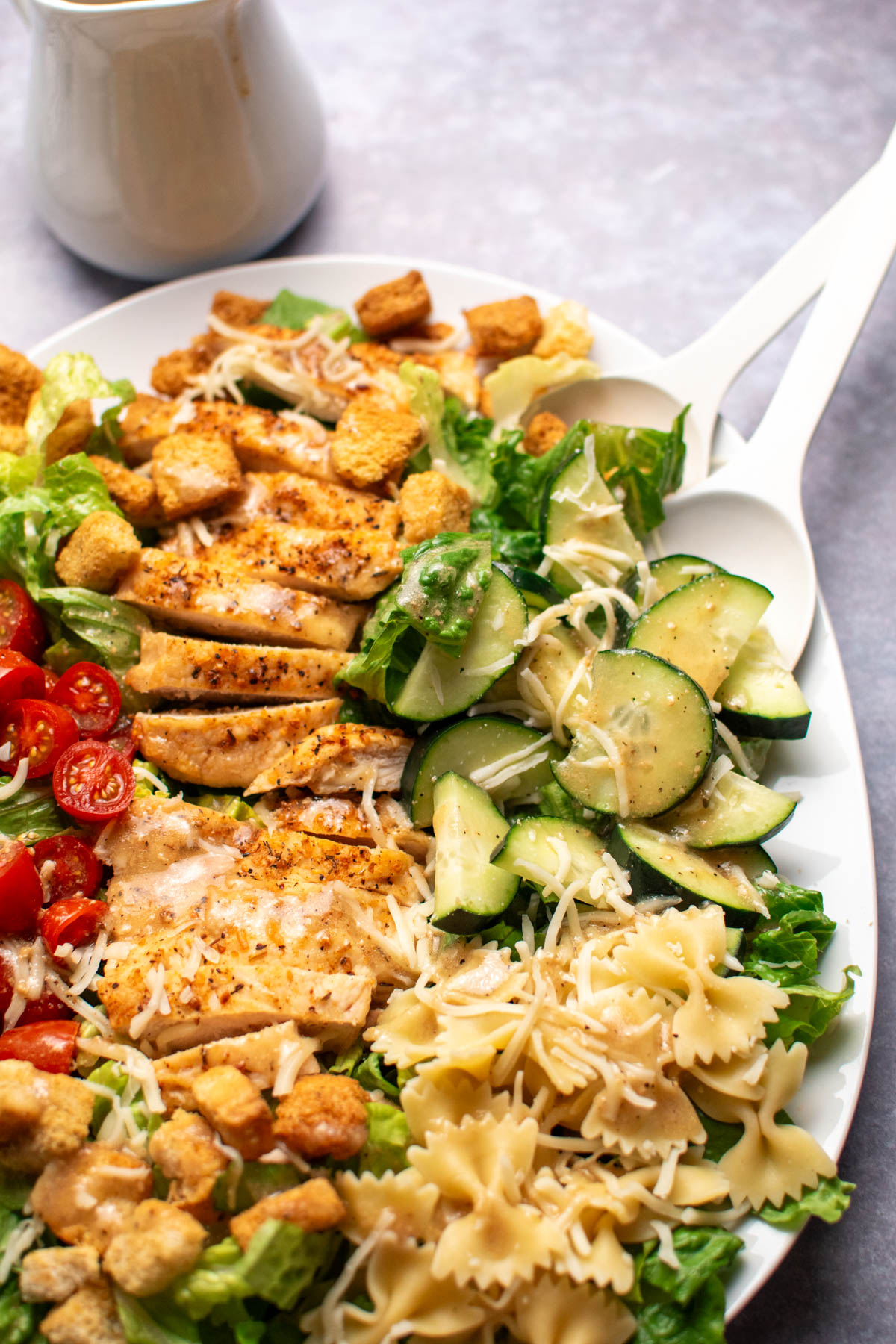 Two salad spoons rest in chicken Caesar salad with pasta, cucumbers, and croutons.