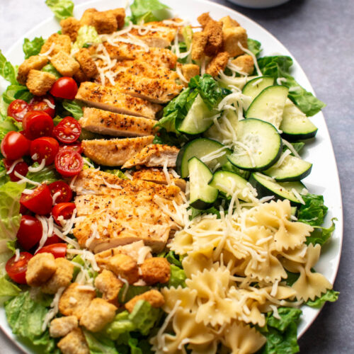 Platter of chicken Caesar pasta salad with tomatoes and cheese on dark table top.