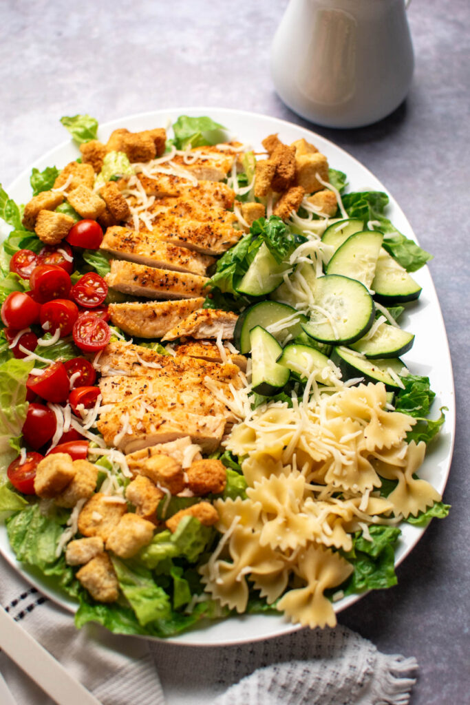 Platter of chicken Caesar pasta salad with tomatoes and cheese on dark table top.