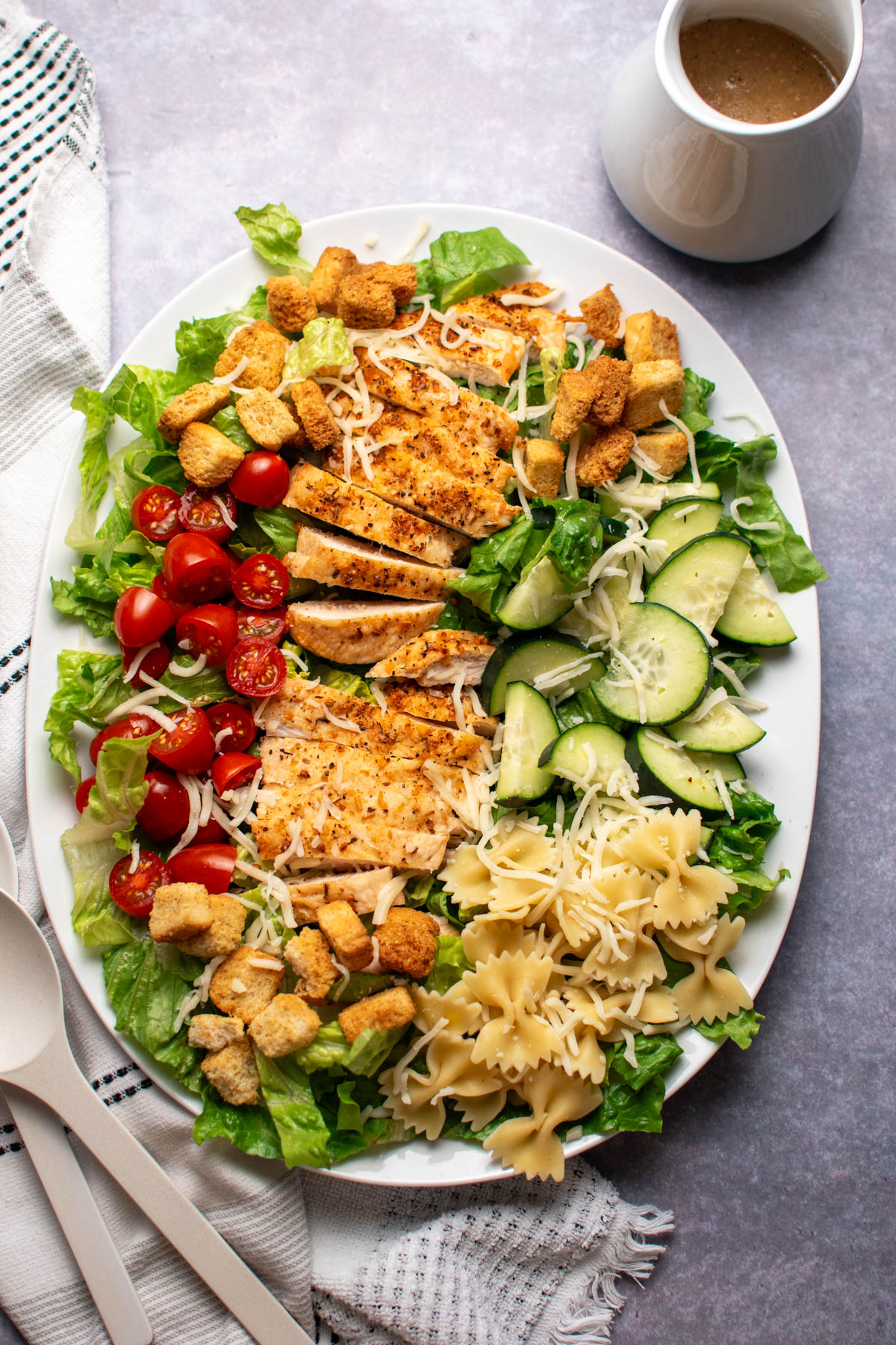 Platter of chicken caesar pasta salad with tomatoes and cucumbers on dark table top with towel and dressing nearby.