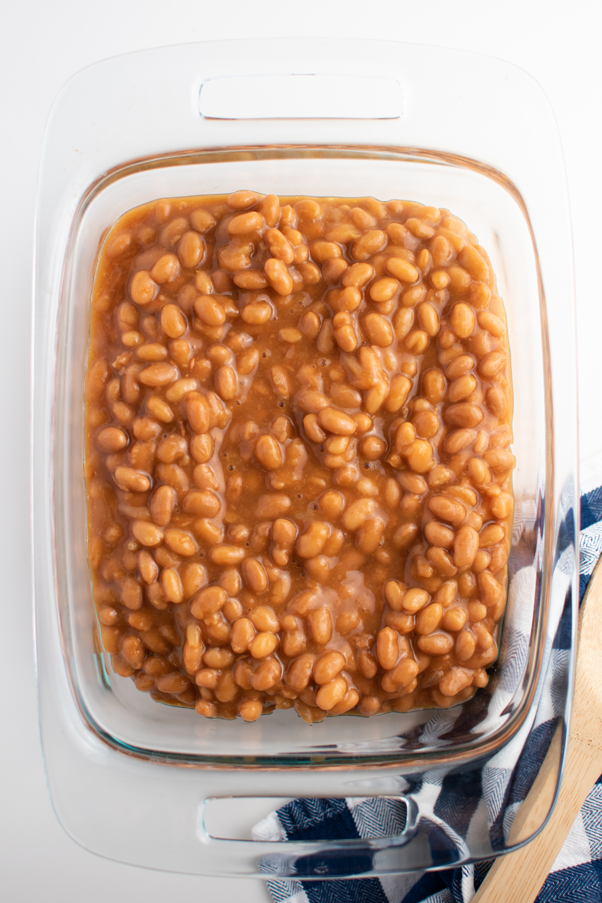 Baked beans in clear glass baking dish next to towel and wooden spoon.