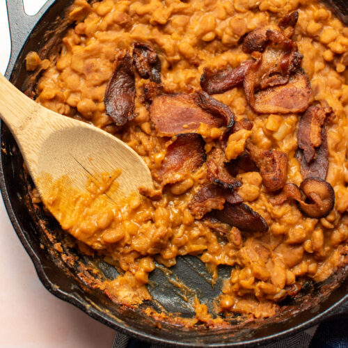 Wooden spoon in cast iron skillet of baked beans with brown sugar topped with bacon.