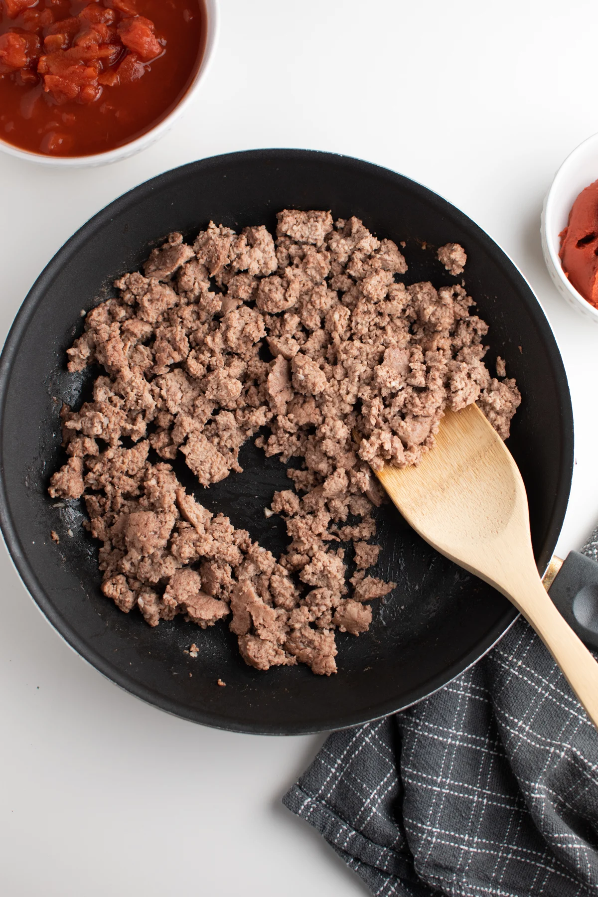 Browned ground beef in black skillet with wooden spoon on white table.