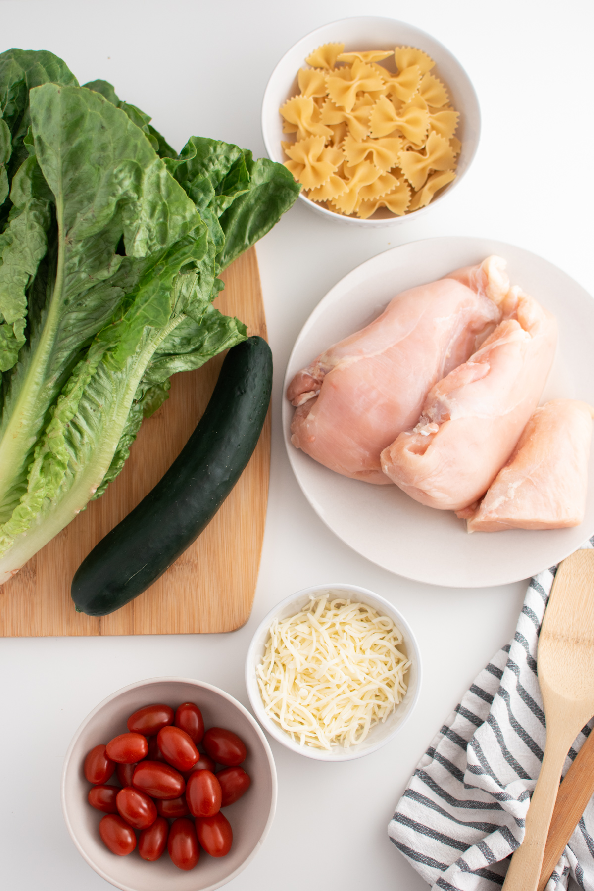 Chicken Caesar salad ingredients including lettuce, cucumber, raw chicken and bowtie pasta.