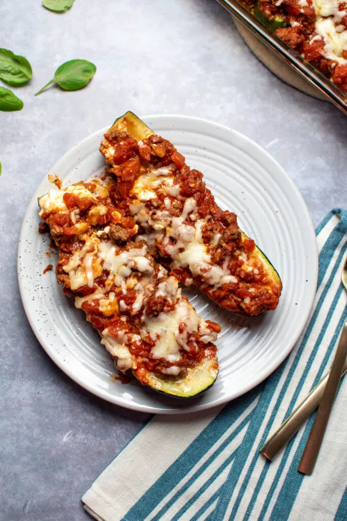 Two lasagna zucchini boats with cottage cheese and mozzarella on a plate surrounded by a kitchen towel and basil.