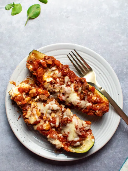 Two lasagna zucchini boats and gold fork on a plate with fresh basil leaves scattered nearby.