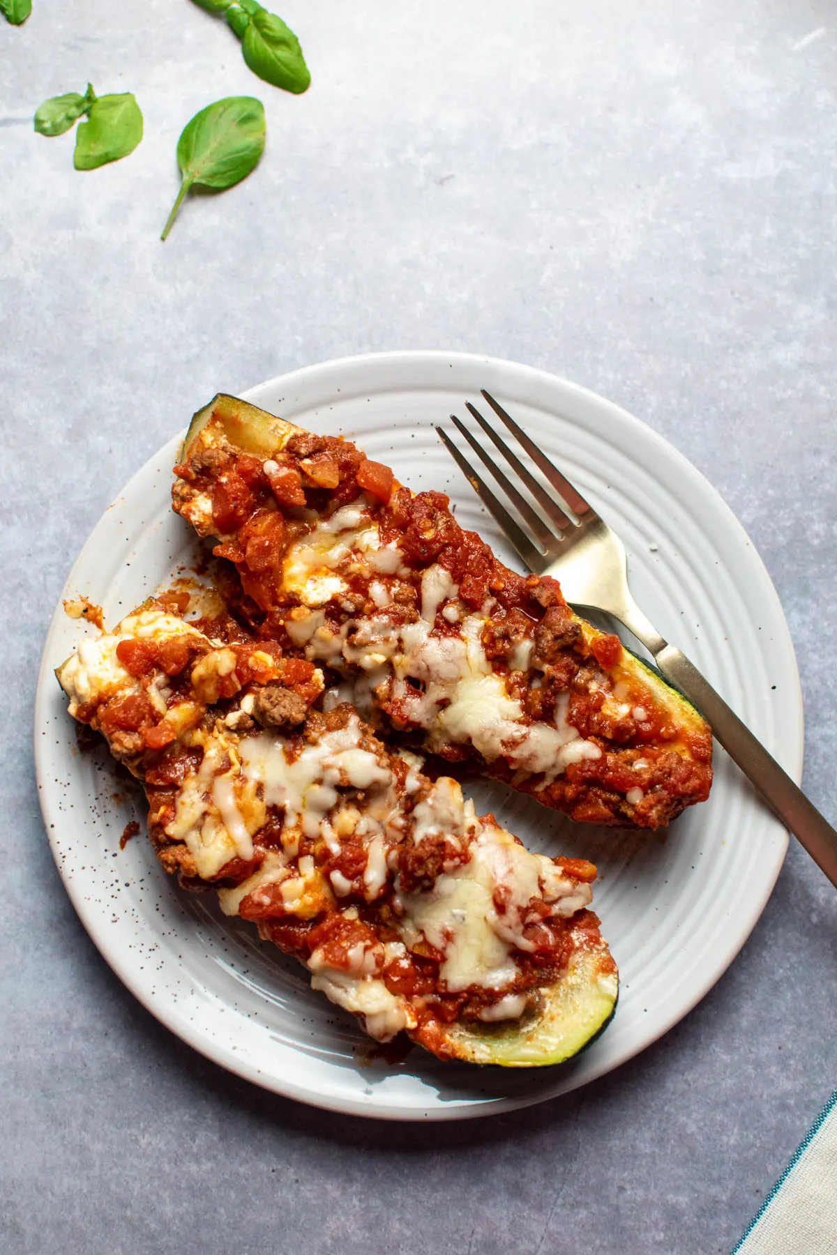Two lasagna zucchini boats and gold fork on a plate with fresh basil leaves scattered nearby.