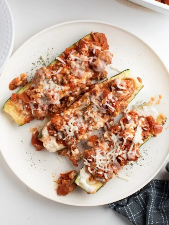 Three lasagna zucchini boats on a white plate with blue kitchen towel.