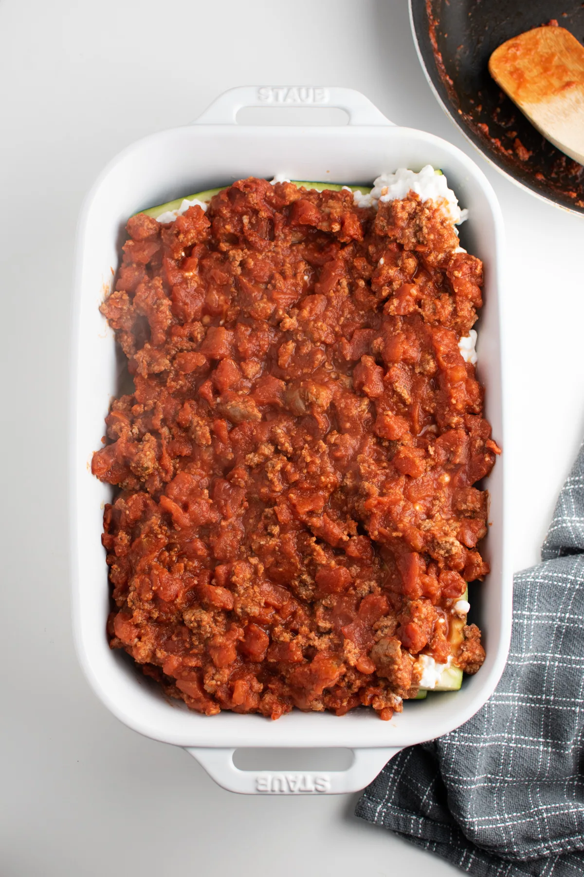 Lasagna meat sauce covering zucchini boats with cottage cheese in white baking dish.