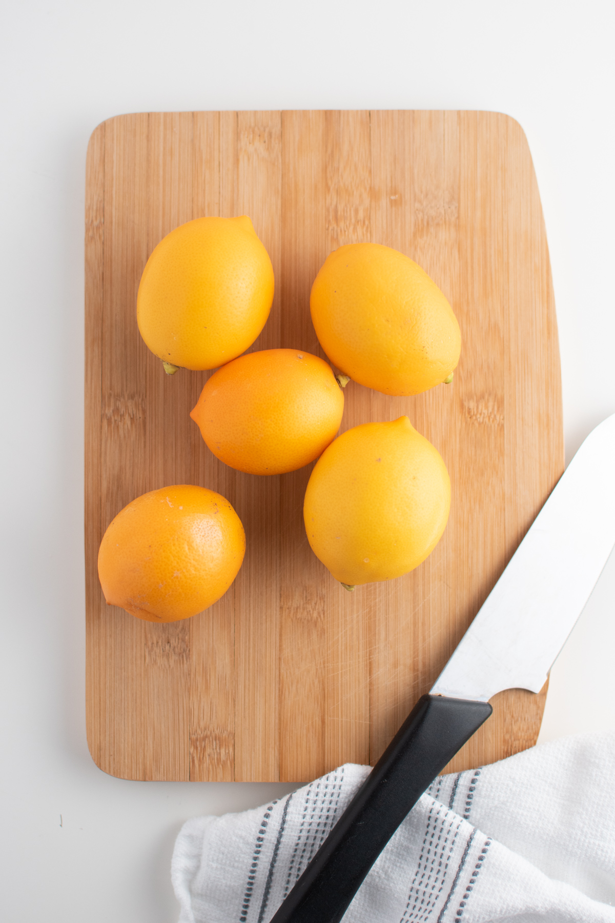 Five Meyer lemons on wooden cutting board with kitchen knife and towel.