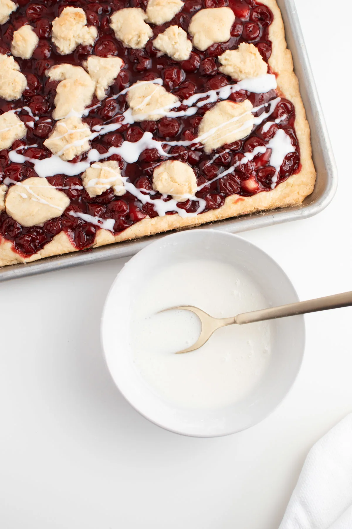 Powdered sugar glaze in white bowl next to sheet pan pie with cherries.