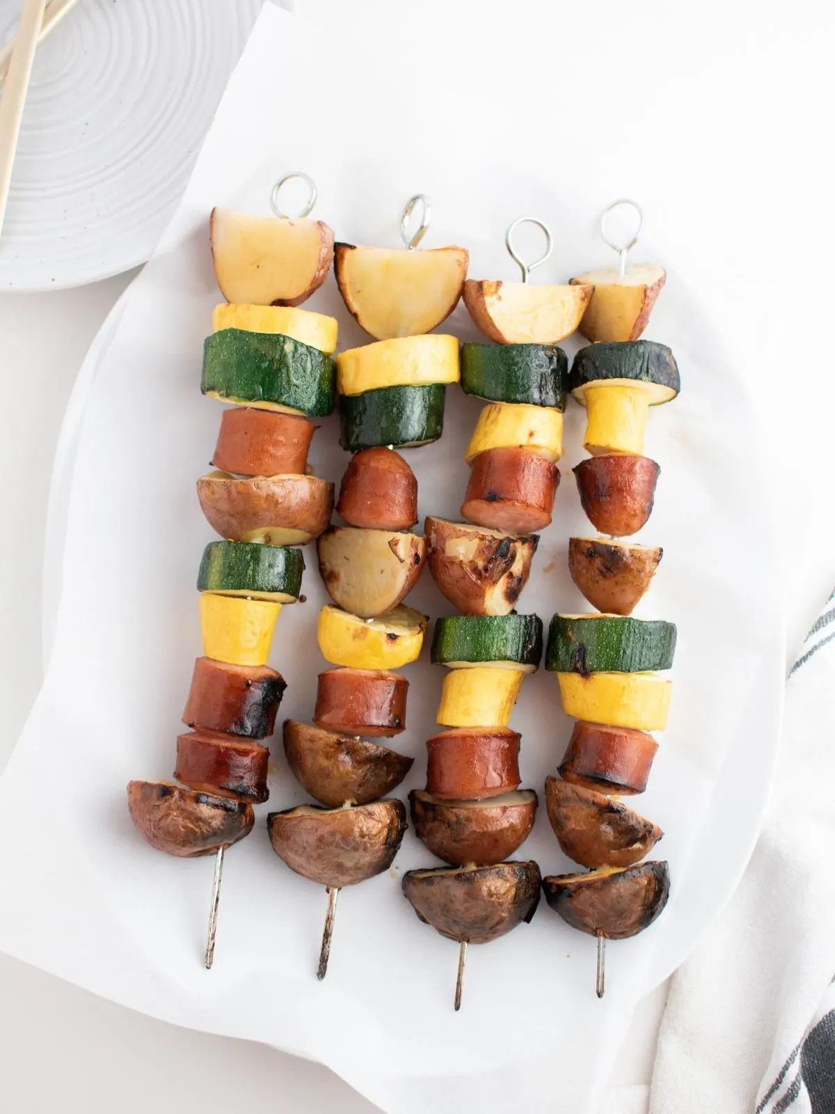 Grilled sausage kabobs with yellow squash, zucchini, and potatoes on a white plate.