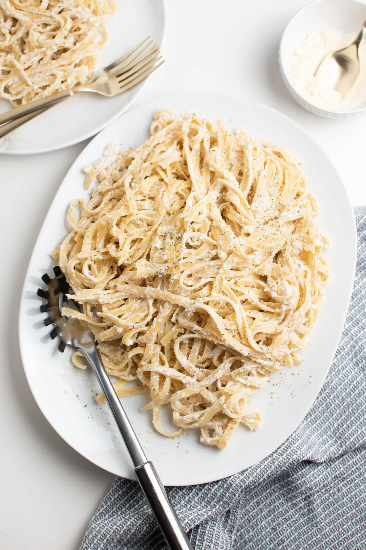 Skinny fettuccine alfredo on white platter with metal slotted spoon and kitchen towel.