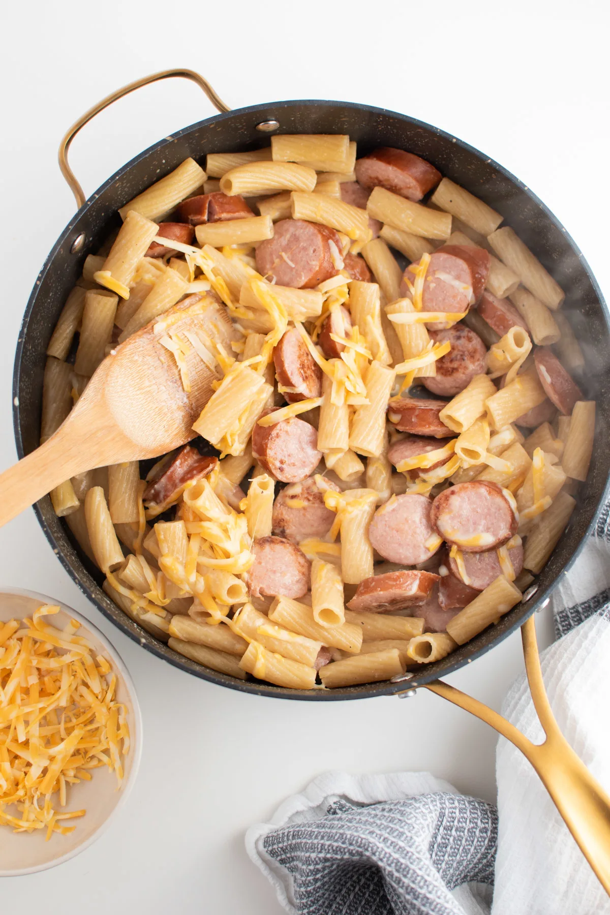 Shredded cheese on top of steamy kielbasa pasta in skillet with wooden spoon.