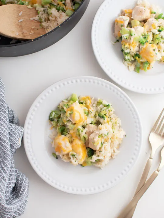 Two white plates with cheesy chicken broccoli and rice surrounded by towel, utensils, and large skillet.
