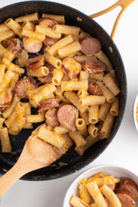 Cheesy kielbasa pasta in large black skillet with wooden spoon on white table.