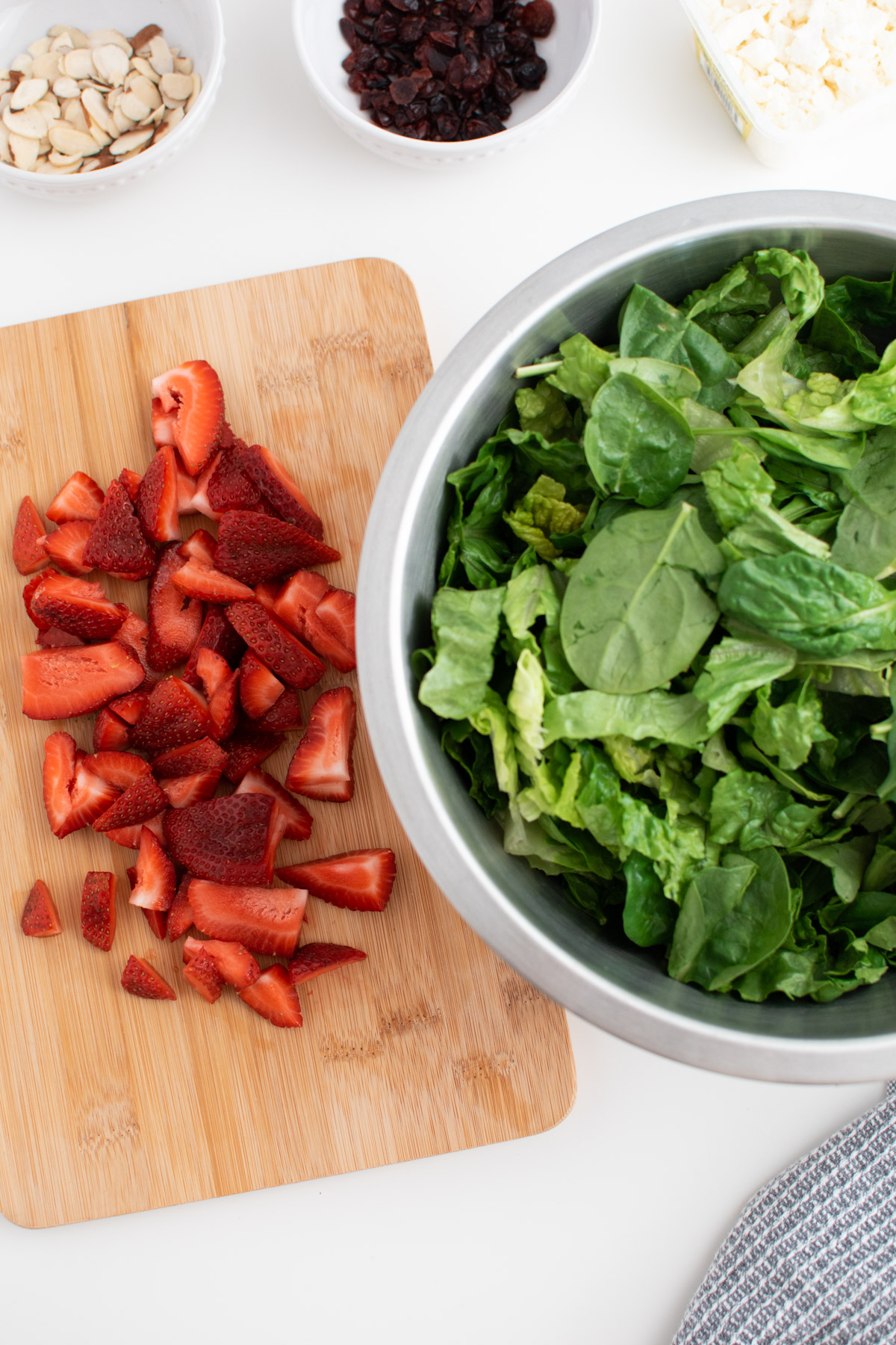 Chopped strawberries on wooden cutting board next to mixing bowl filled with lettuce and spinach.