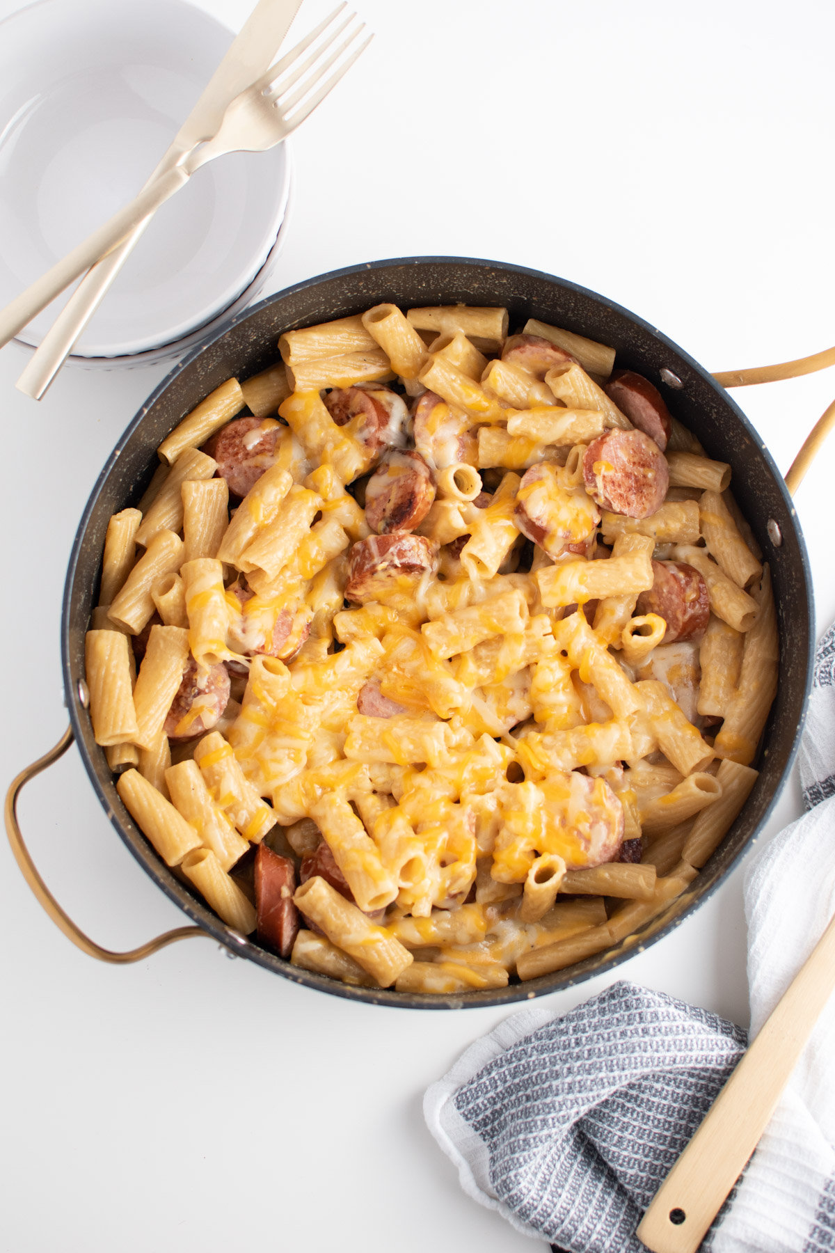 Kielbasa rigatoni pasta with melted cheese in large skillet surrounding by stack of plates and towel.