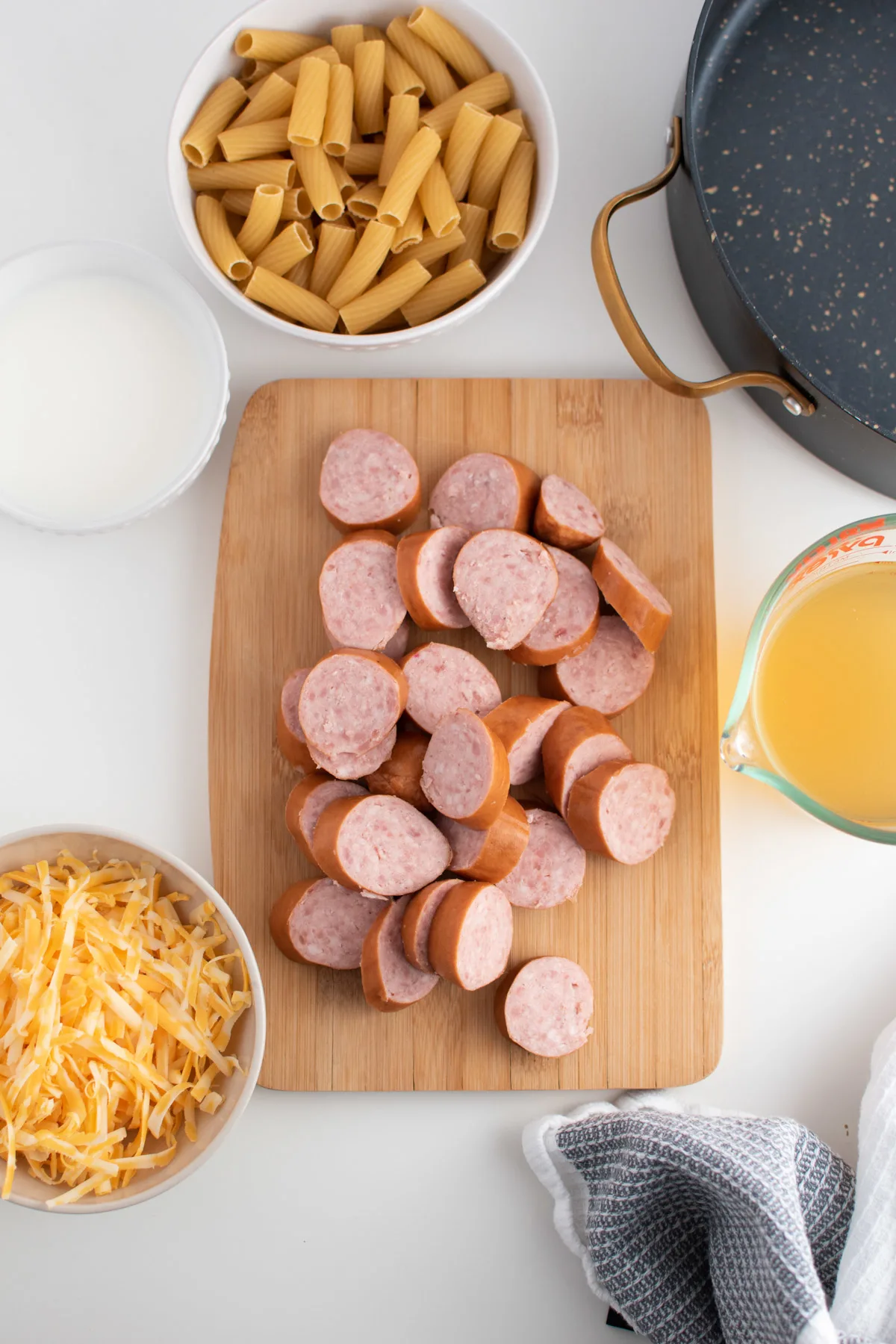 Kielbasa pasta ingredients including shredded cheese, milk, broth and sausage on white table.