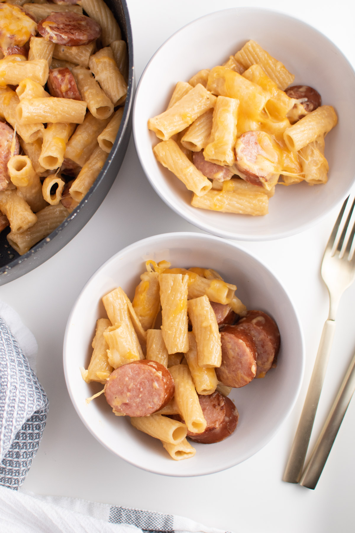 Two white bowls filled with kielbasa pasta next to gold utensils, skillet, and towel.