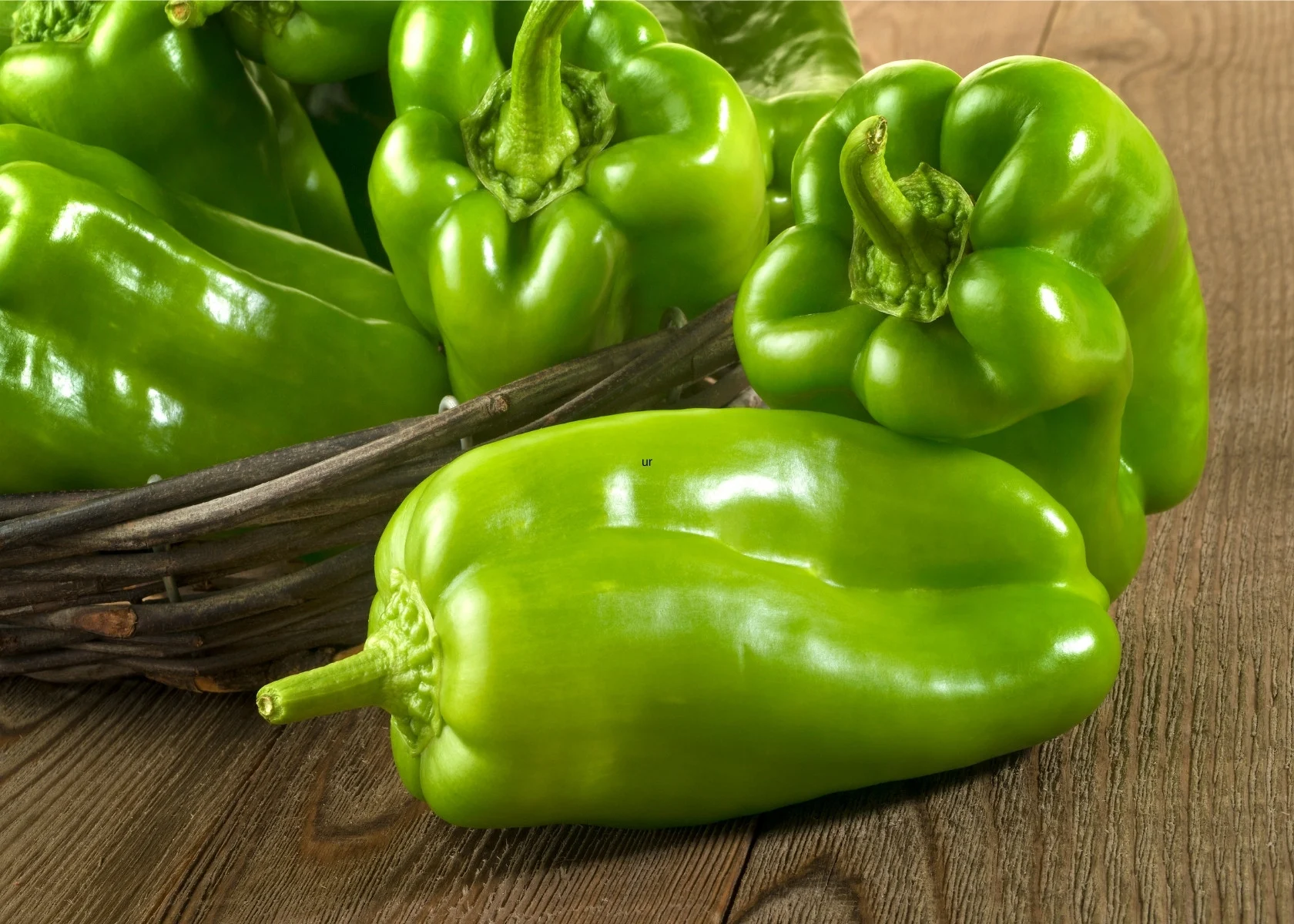Several bright green bell peppers spilling from whicker basket onto wooden table.