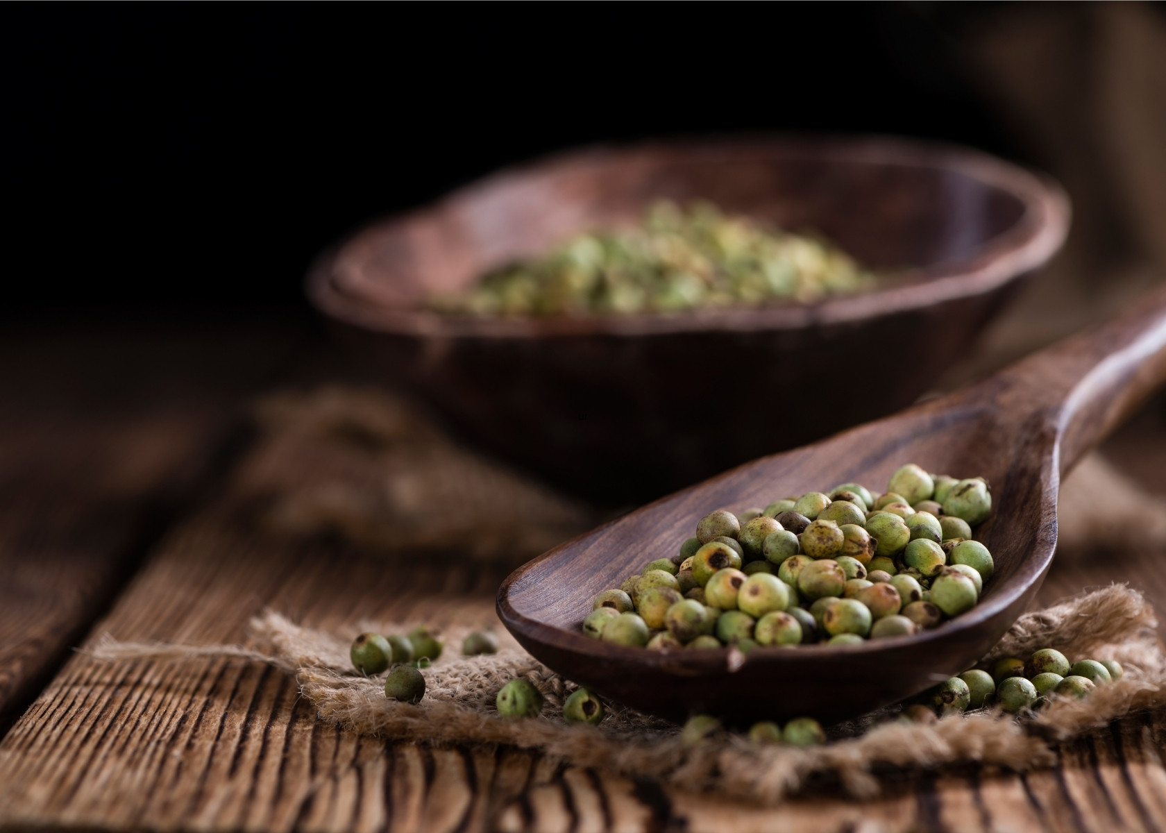 Green peppercorns in wood bowl next to large wooden spilled filled with more peppercorns.