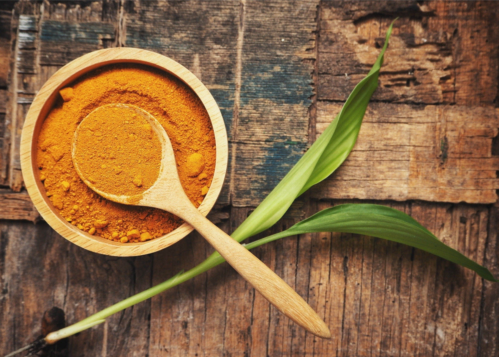 Bright orange ground turmeric in wooden bowl with large wooden spoon with green leaf.
