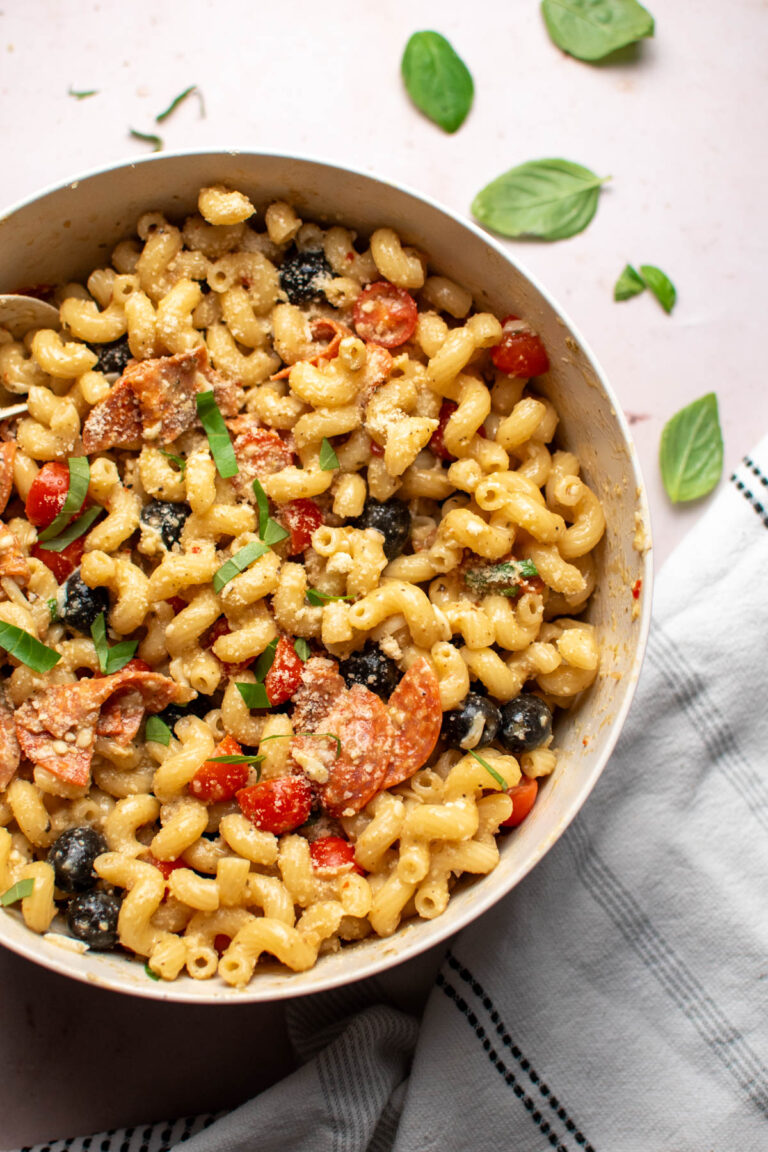 Pizza pasta salad in large tan bowl with fresh basil leaves on table top.