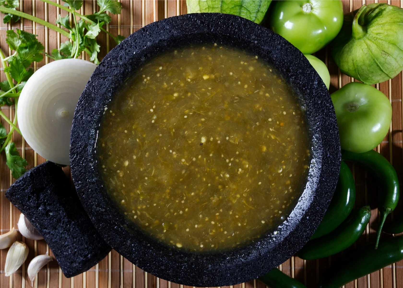 Salsa verde in large dark colored mortar and pestle next to fresh tomatillos and onion.