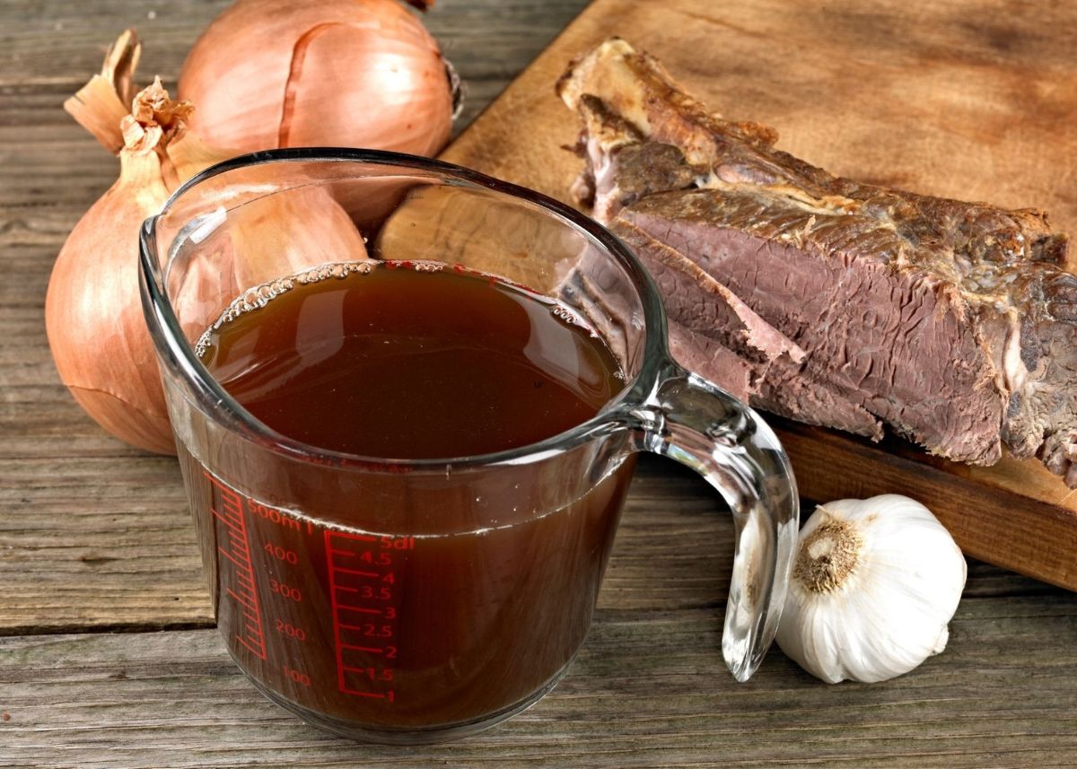Beef broth in glass measuring cup next to onions, garlic and slab of beef.