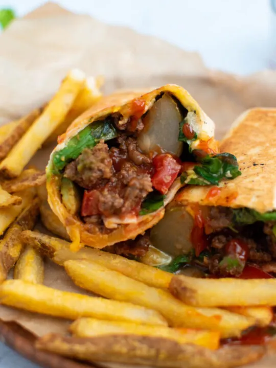 Grilled cheeseburger wrap cut in half surrounded by French fries on wood plate.