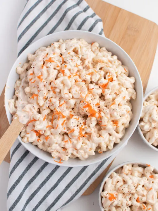 Bowl of Hawaiian macaroni salad with carrots on wooden cutting board next to kitchen towel.