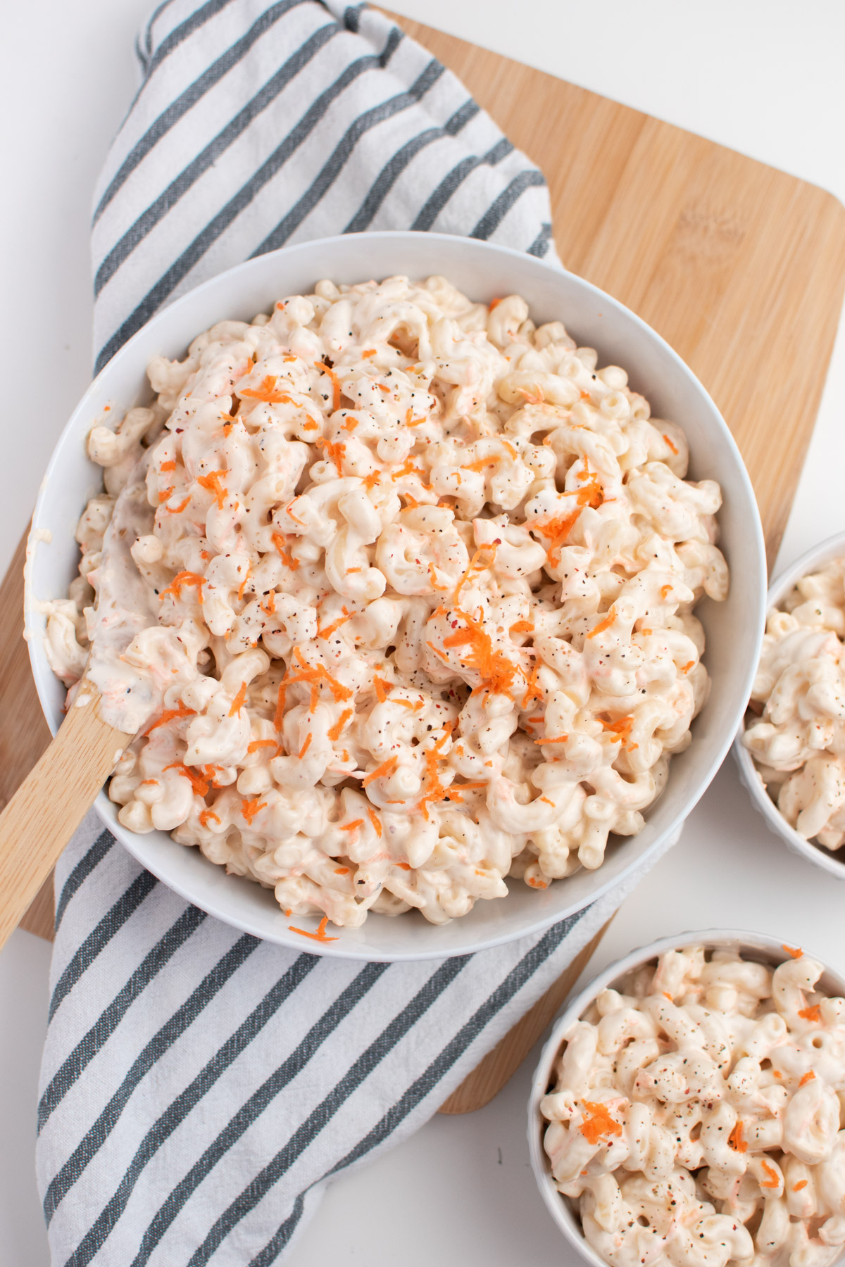 Bowl of Hawaiian macaroni salad with carrots on wooden cutting board next to kitchen towel.