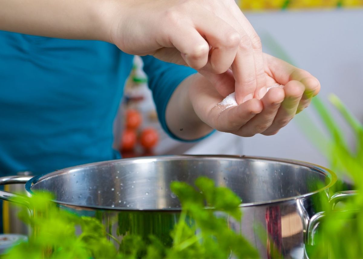 Woman holds salt in her palm over large metal stock pot of water next to fresh herbs.