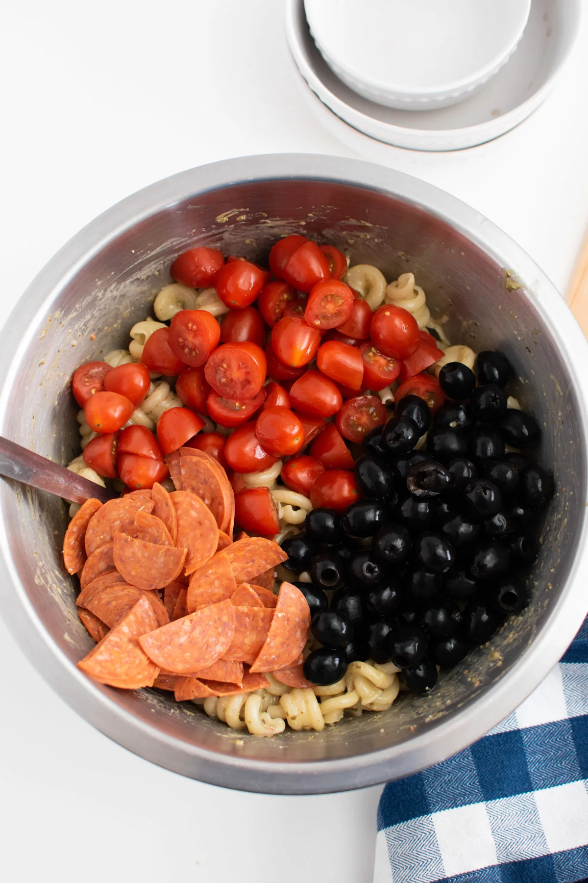 Tomatoes, olives, and pepperoni on top of pasta in large metal bowl.