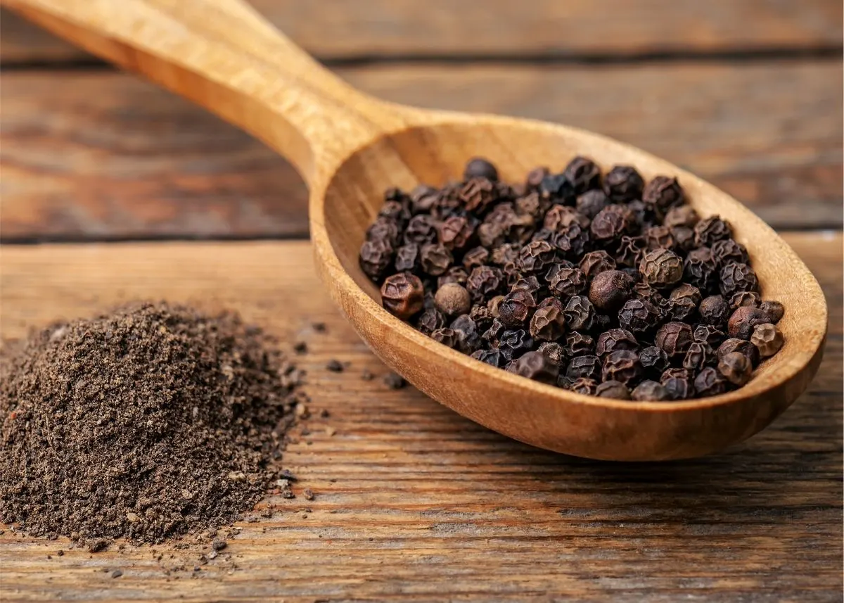 Lots of black peppercorns on a wooden spoon next to pile of ground black pepper.