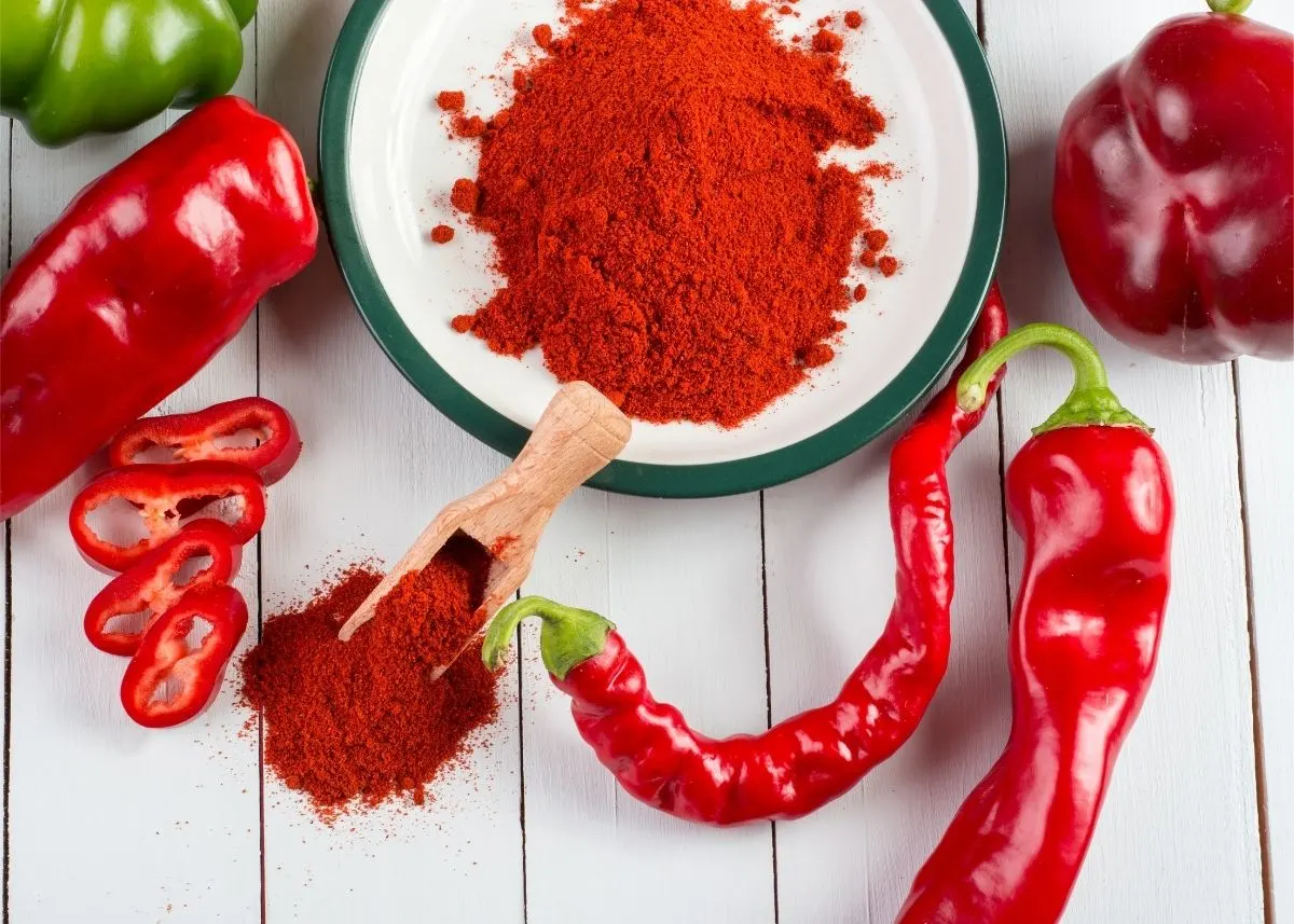 Large mound of reddish-orange paprika powder in bowl next to assortment of fresh red chilis.