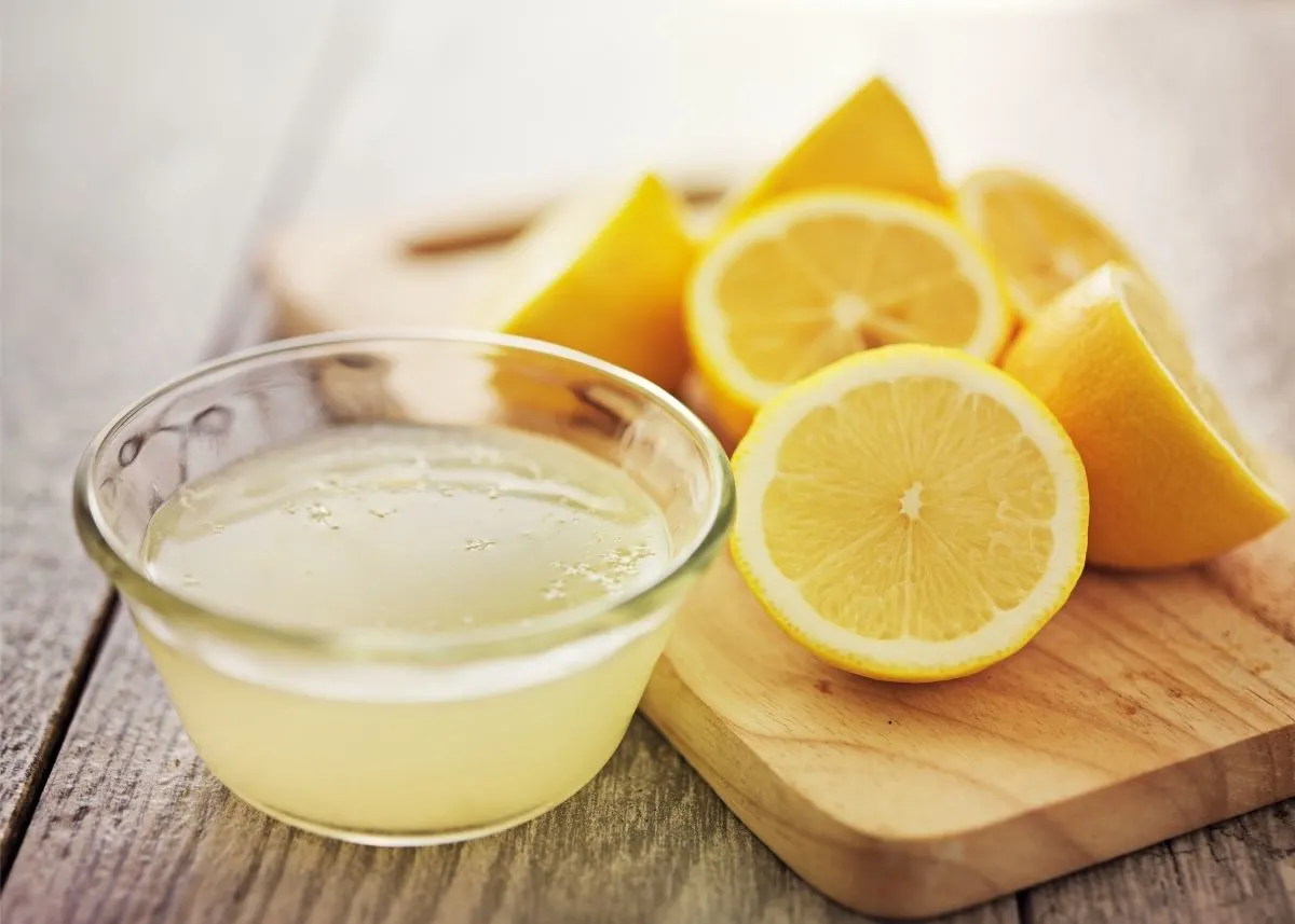 Several cut lemons on a wooden cutting board next to a bowl of lemon juice.