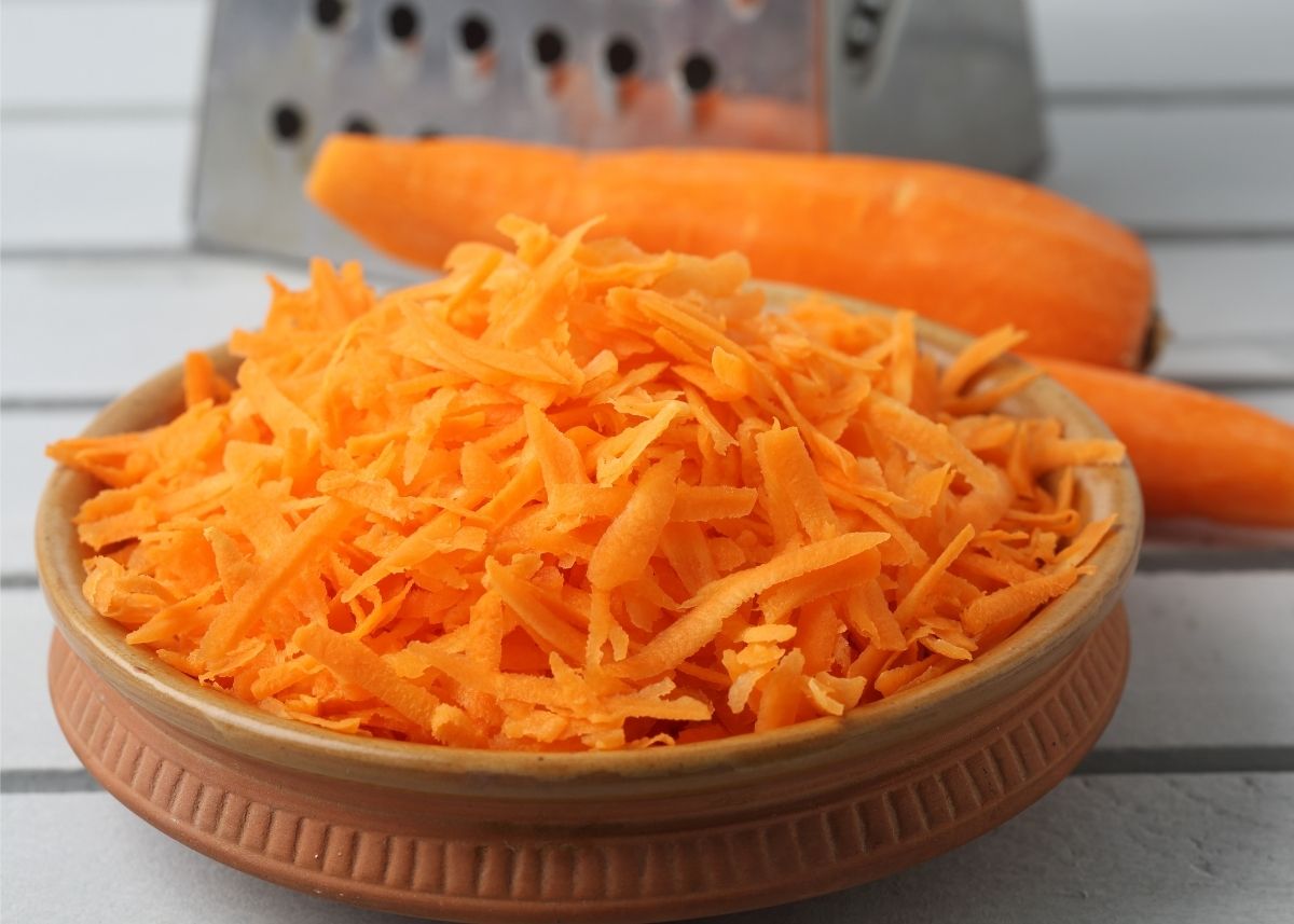 Metal kitchen grater sits behind whole carrot stalk and bowl of shredded carrot.
