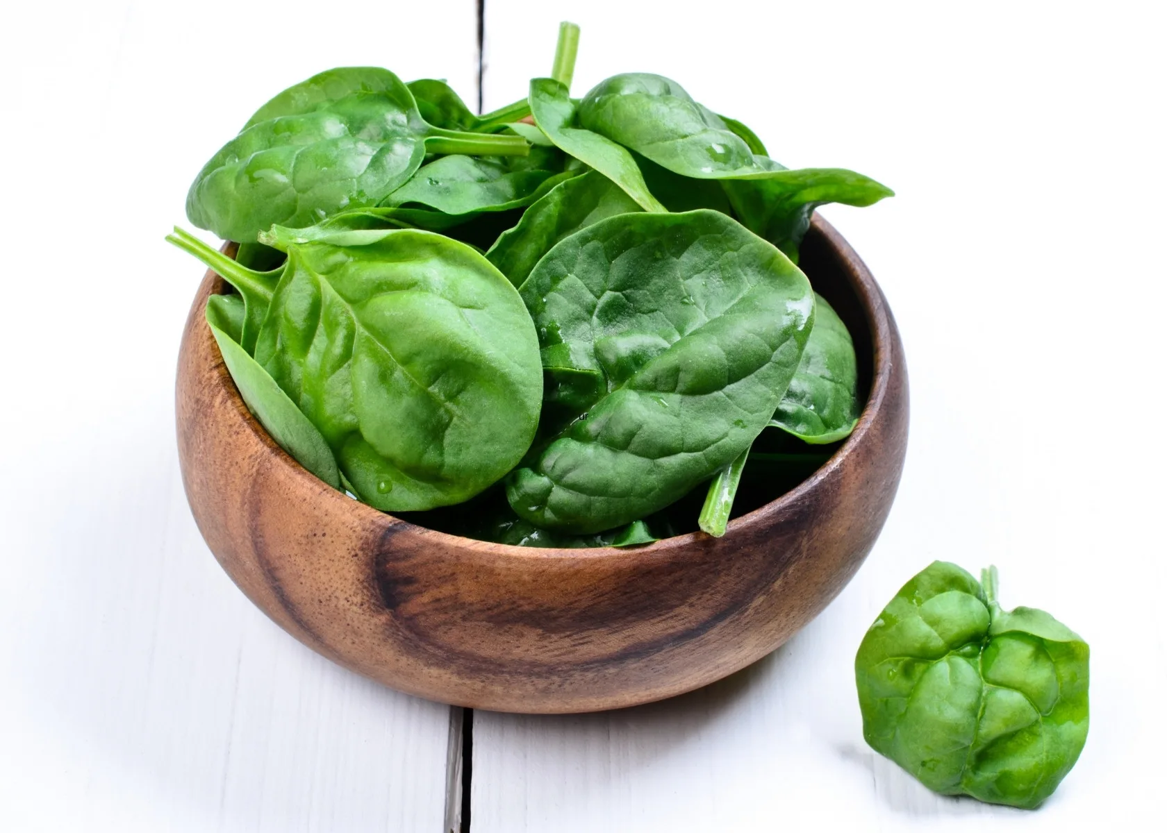 Freshly washed large spinach leaves in dark wooden bowl on bright white kitchen table.