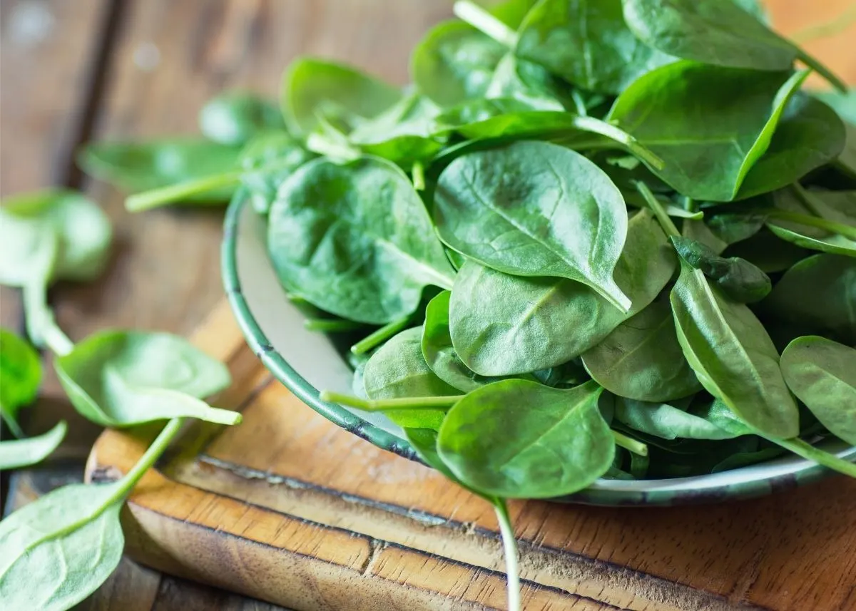 Spinach leaves piled in a shallow bowl on top of a rustic wooden table.