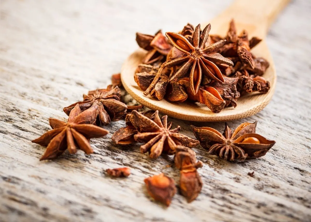 Large pile of star anise spilling off of a wooden spoon on wooden table top.