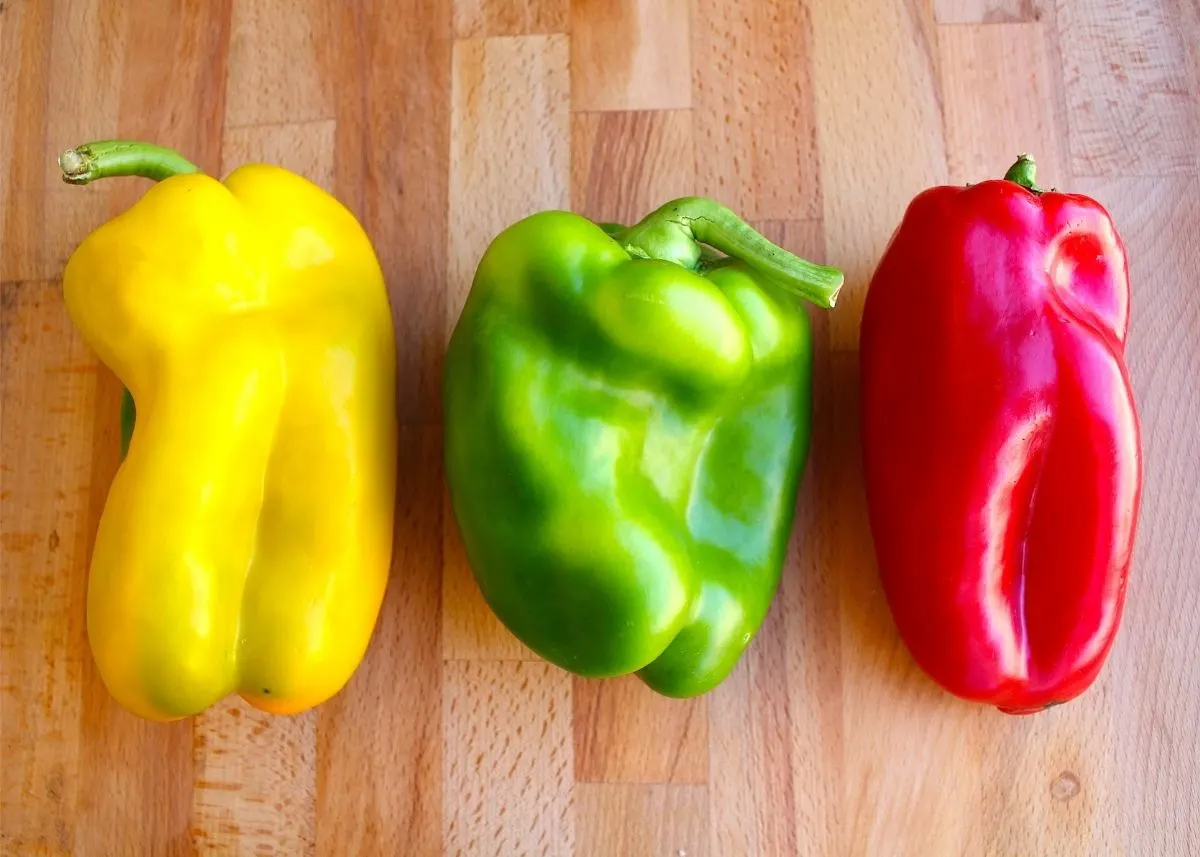 Trio of red, green and yellow bell peppers lined up on a wooden cutting board.