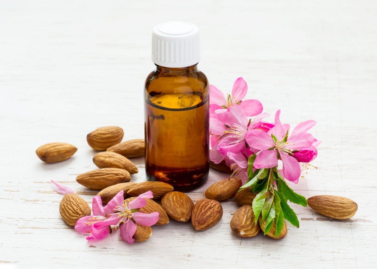 Bottle of almond essential oil surrounded by scattered almonds and pink almond flowers.