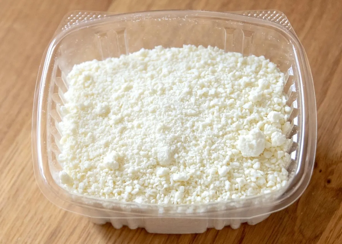 Large amount of Cotija Molido cheese in a clear plastic deli container on wood table.