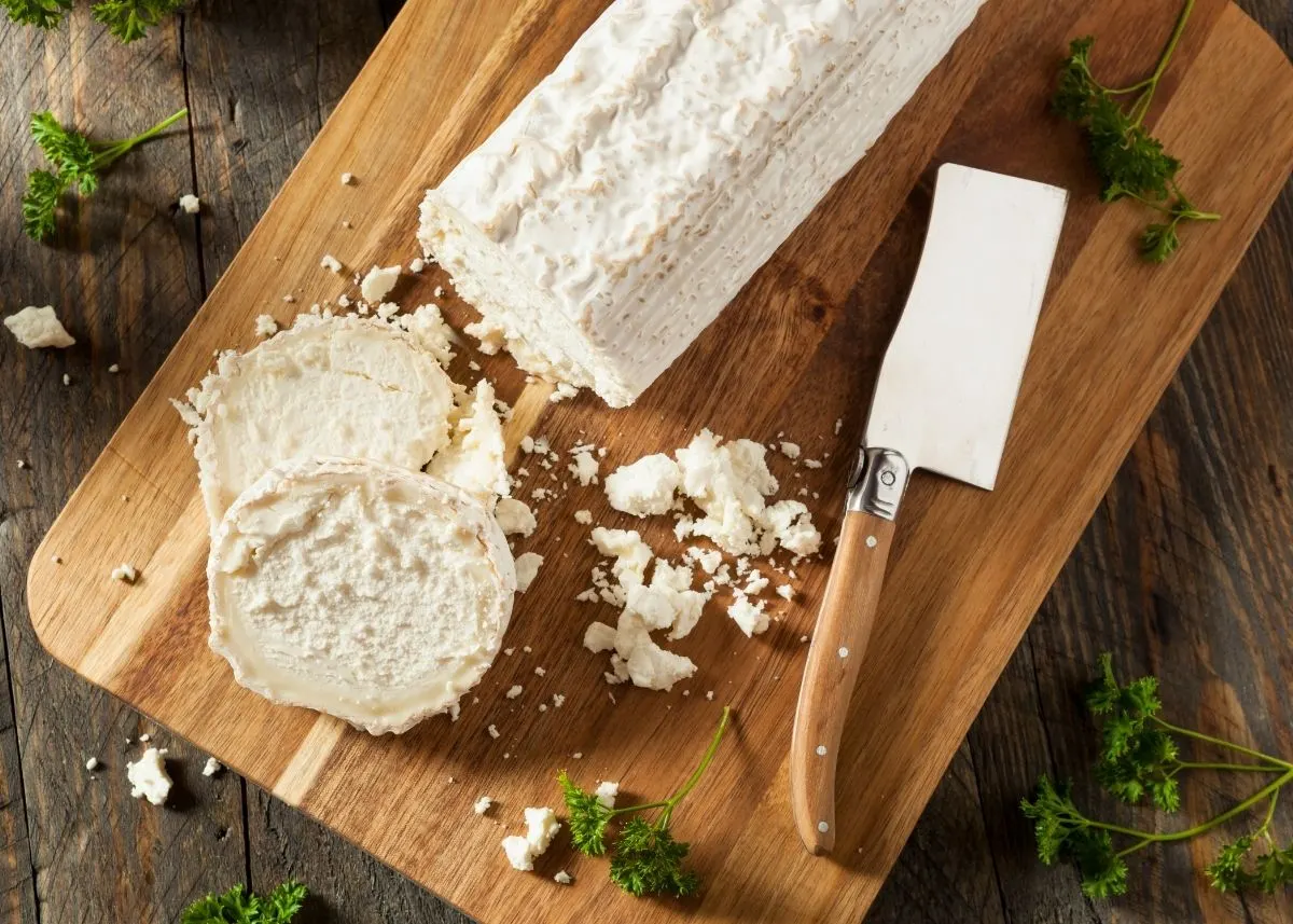 Sliced goat cheese log and goat cheese crumbles on a wooden cutting board next to small cleaver.