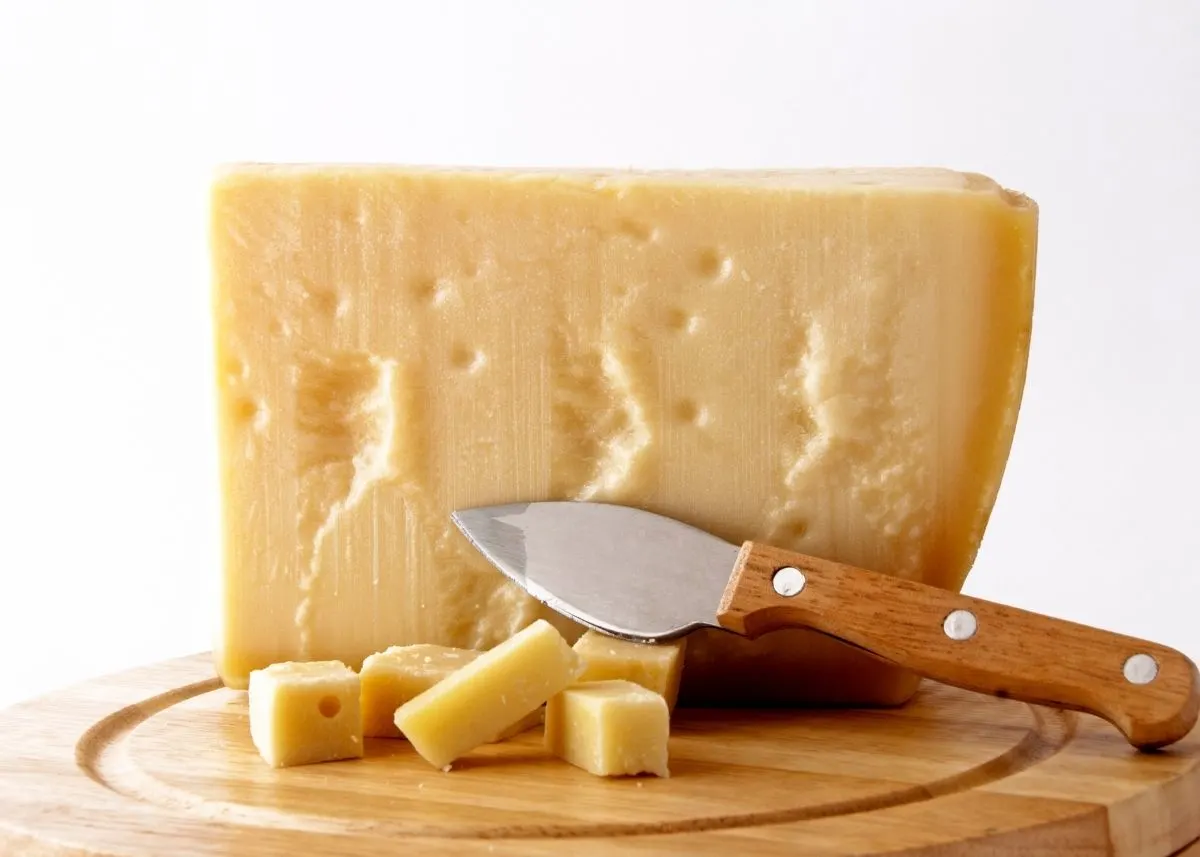 Large portion of a Grana Padano cheese wheel next to slices and cheese knife on a cutting board.