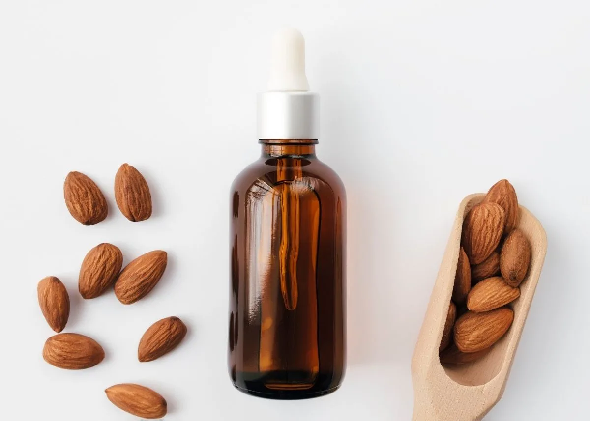 Brown bottle of imitation almond extract next to a wooden spoon and pile of almonds.